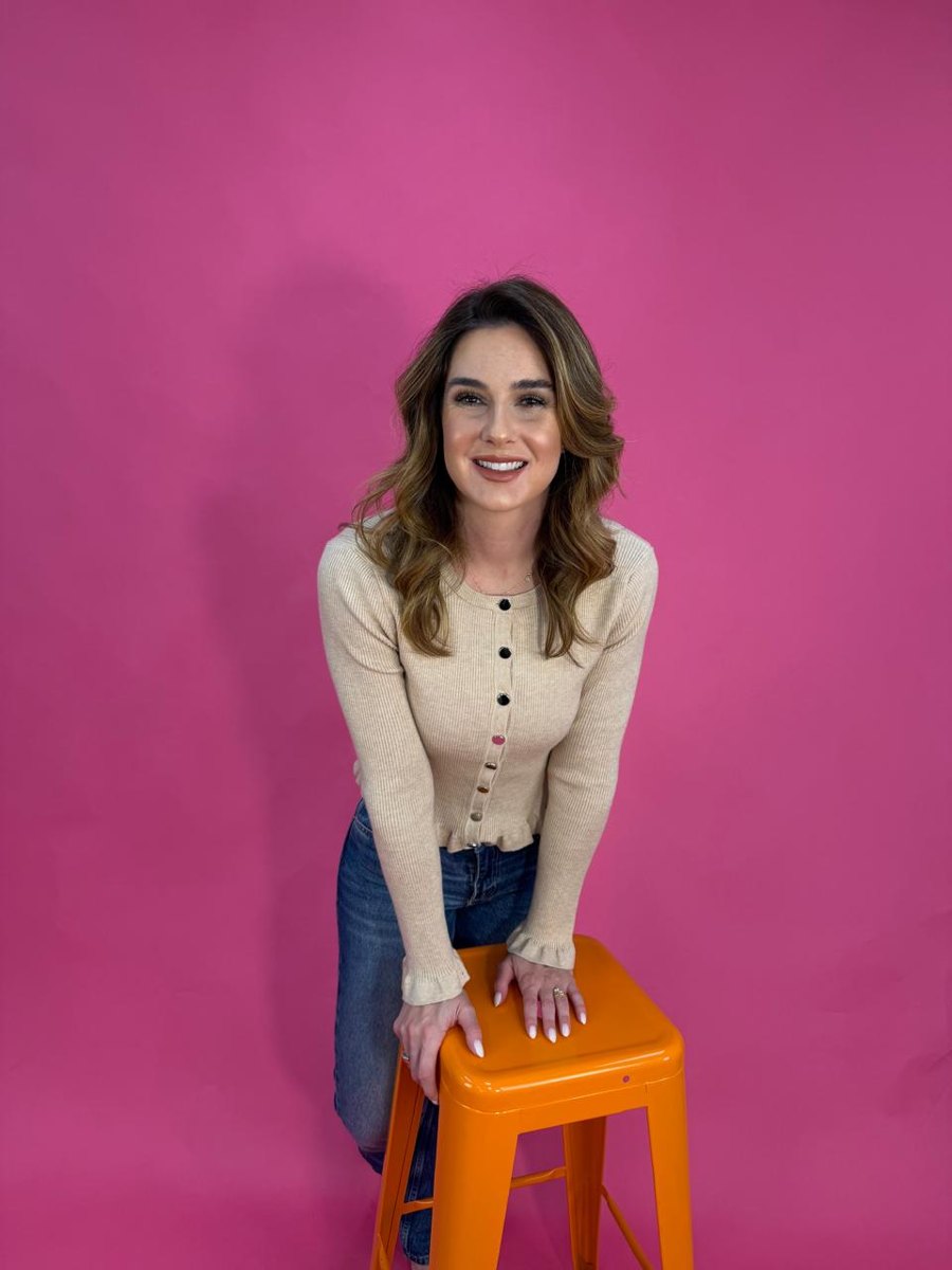 Aline sorrindo, apoiada em um banco laranja diante de um fundo rosa